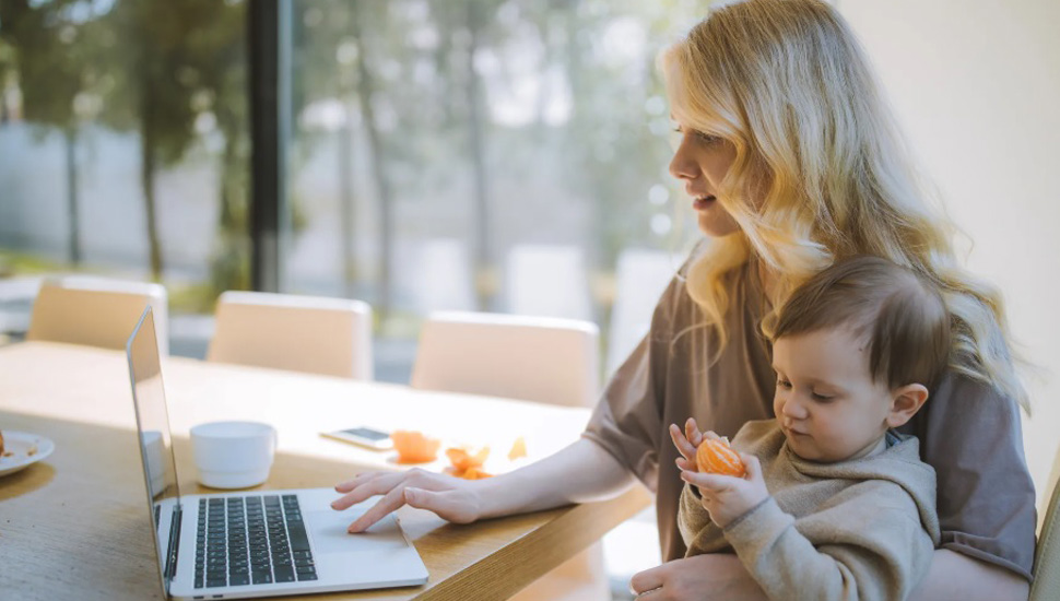 Žena radi na laptopu dok drži dete u krilu