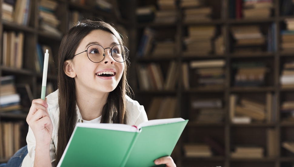 Devojčica u biblioteci drži oduševljeno svesku i olovku