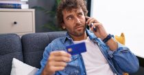 young-hispanic-man-talking-smartphone-holding-credit-card-sitting-sofa-home